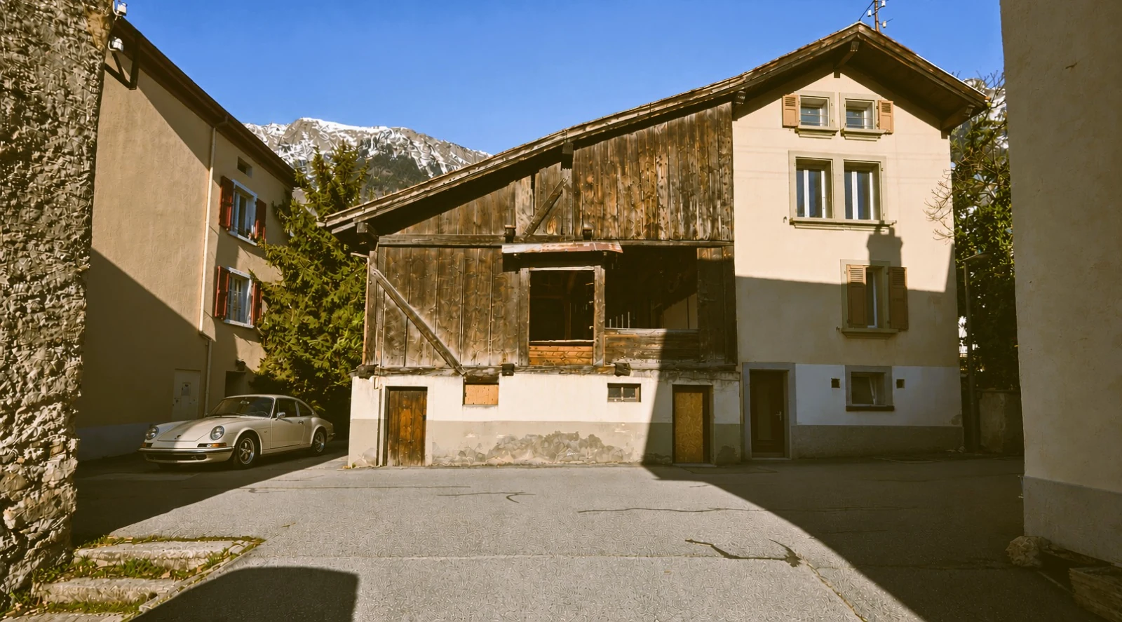 La grange en planches de bois sombre et la maison enduite, vues depuis la cour : la grange ouverte de deux grandes baies au-dessus d’un socle enduit percé de portes, la maison à volets de bois ; à gauche, une maison voisine et une voiture ancienne garée, au fond les montagnes enneigées