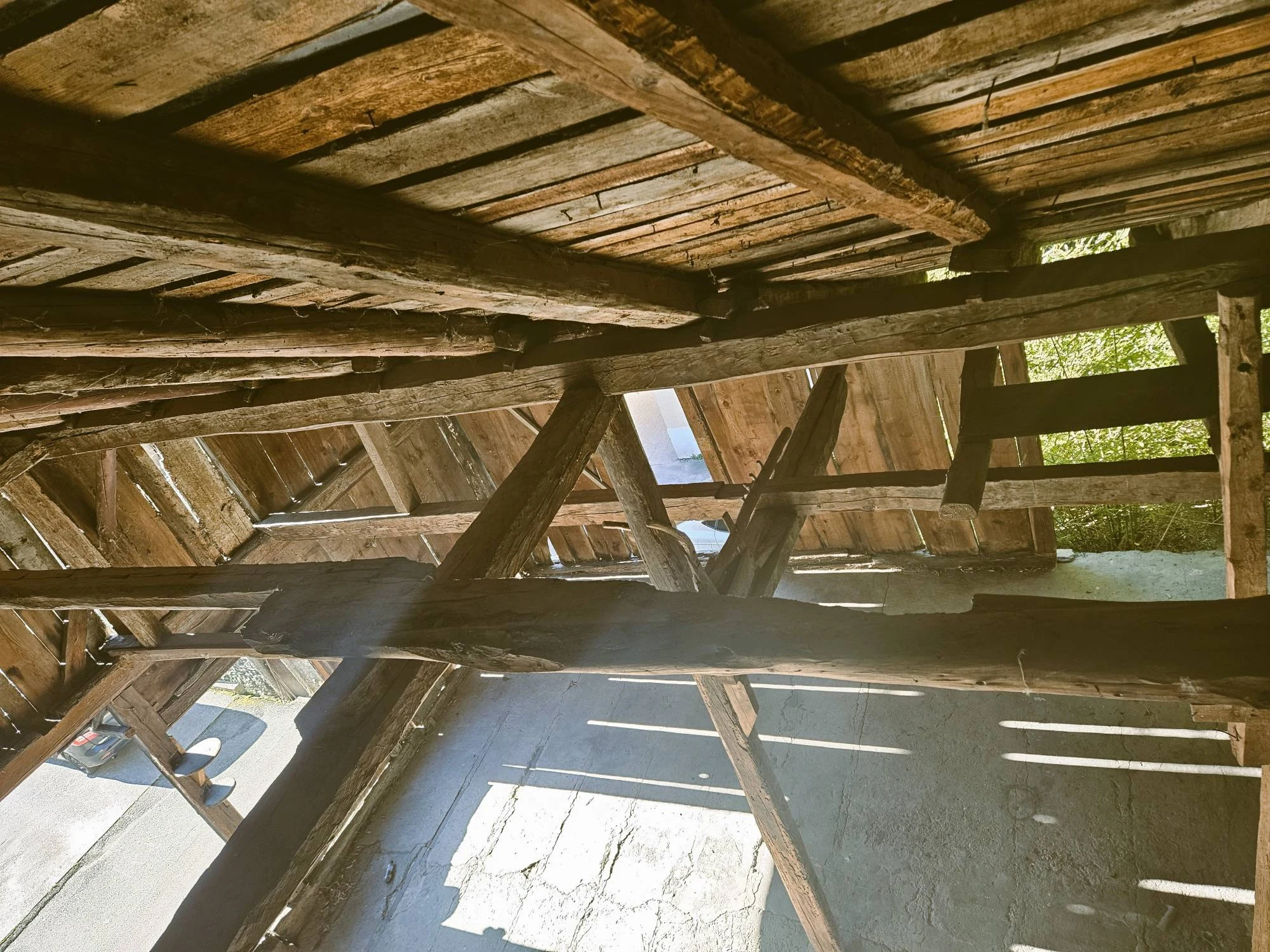 Sous un plancher de planches porté par de grosses poutres, des contrefiches de bois ; le jour passe entre les planches du bardage et dessine des rayures sur la dalle ; à gauche, une ouverture sur la rue