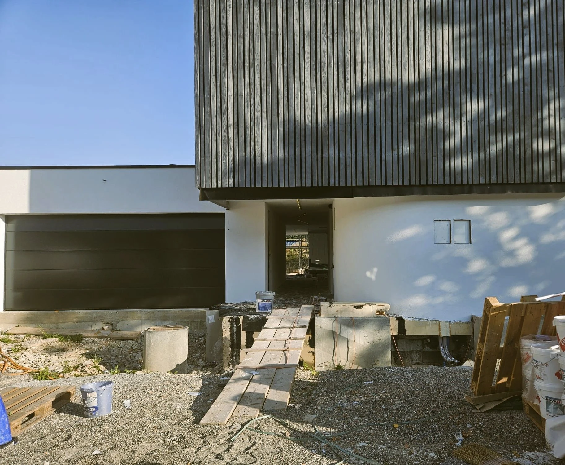 Le garage à porte sombre à gauche, le passage couvert au centre, ouvert de part en part, et au-dessus le volume de l’étage en bardage de bois sombre ; une passerelle de planches mène au passage